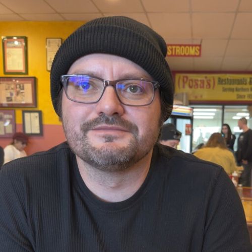 Randy Davila in a restaurant, he is wearing a black knit cap and black shirt and glasses