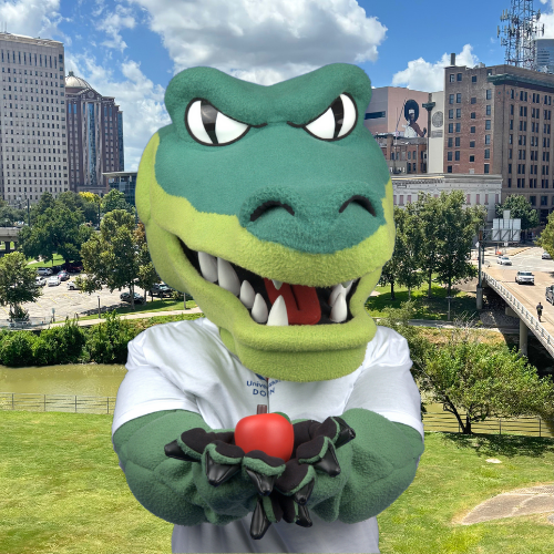 university of houston downtown mascot Ed u gator holds a red apple in front of the bayou and downtown houston skyline