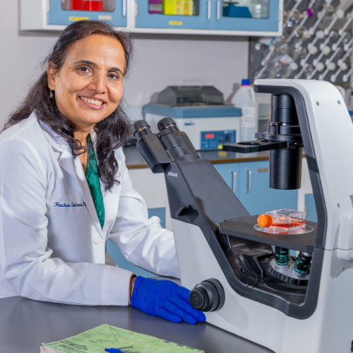 Rachna Sadana wears a lab coat and gloves while operating a microscope in a lab at the university of houston downtown