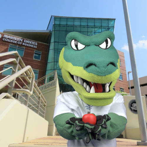 ed u gator university of houston downtown mascot holds an apple while standing in front of marilyn davies college of business