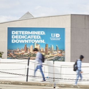 Students walking on a bridge. Sign behind them reads determined, dedicated, downtown and the university of houston downtown logo. 